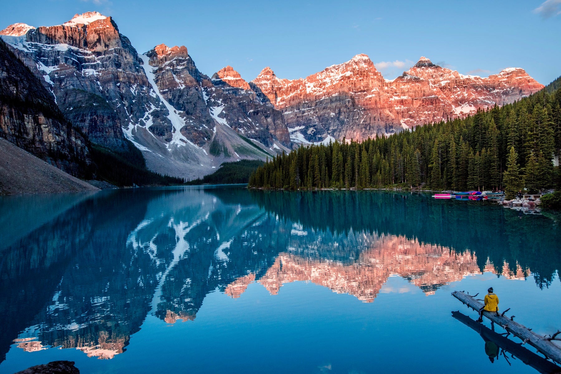 Beautiful outdoor photo of a man sitting on a log overlooking a beautiful lake surrounded by mountains.